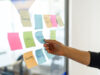 eine Hand greift nach einem Post-It "Metrik" an einer Post-It-Wand