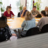 9 Mitarbeiterinnen und Mitarbeiter von MAI eresult im Konferenzraum am großen Besprechungsraum sitzend und diskutierend. Von draußen durch die Glastüre fotografiert.